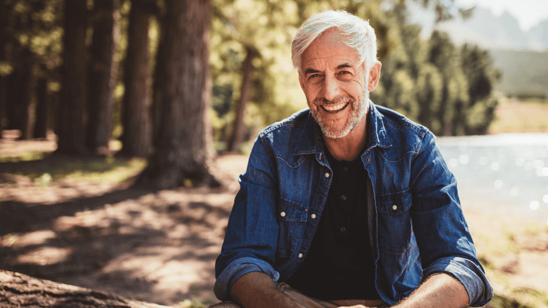Smiling man in denim jacket relaxing by trees near water